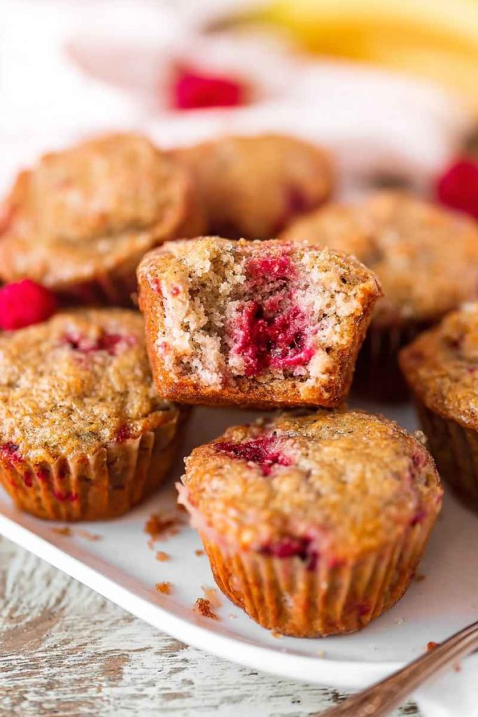 A plate of golden-brown banana raspberry muffins with one halved to reveal a juicy raspberry filling.