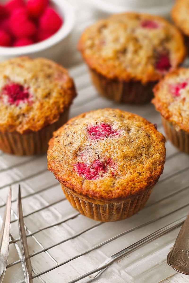 Golden-brown banana raspberry muffins cooling on a wire rack, showcasing their juicy fruit topping.