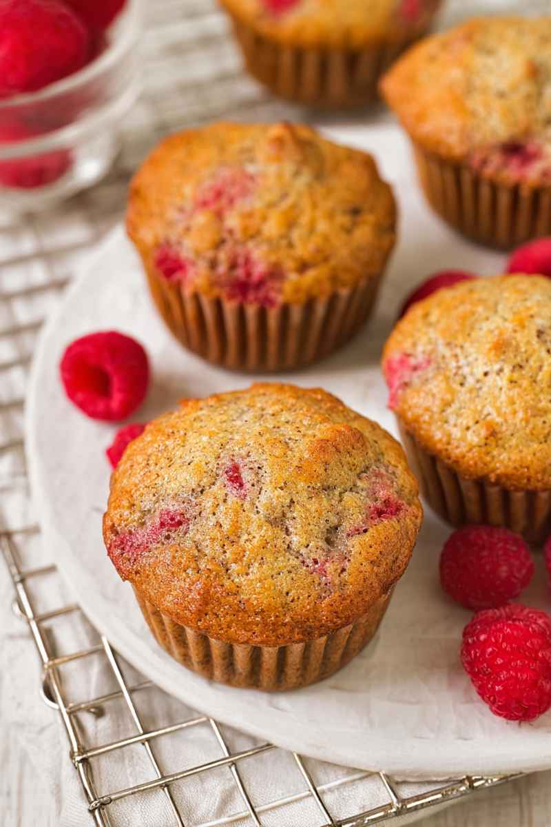 Freshly baked banana raspberry muffins surrounded by vibrant red raspberries on a white plate