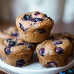A close-up of blueberry muffins stacked on a white plate, with fresh blueberries and a decorative fork nearby.