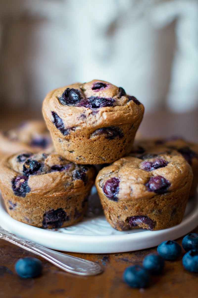A close-up of blueberry muffins stacked on a white plate, with fresh blueberries and a decorative fork nearby.