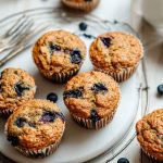 Golden-brown blueberry zucchini muffins with visible blueberries, arranged on a white plate atop a rustic wooden table, surrounded by scattered blueberries and a vintage fork.