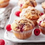 Golden-brown raspberry muffins with a crumbly streusel topping, surrounded by fresh raspberries on a white plate.