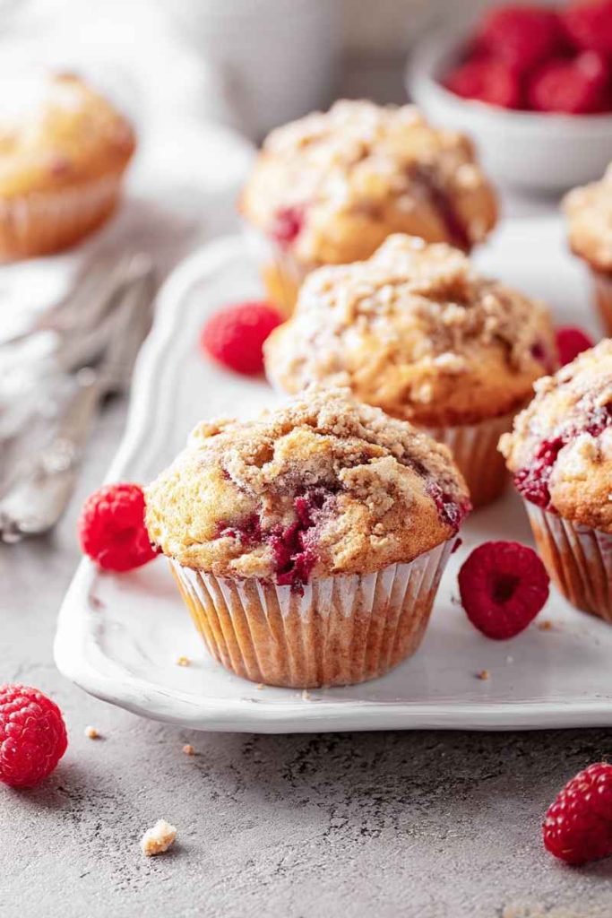 Golden-brown raspberry muffins with a crumbly streusel topping, surrounded by fresh raspberries on a white plate.