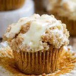 A close-up of a golden-brown carrot cake muffin topped with creamy white frosting and crumbly streusel, sitting on a white plate.