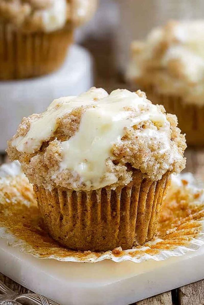 A close-up of a golden-brown carrot cake muffin topped with creamy white frosting and crumbly streusel, sitting on a white plate.