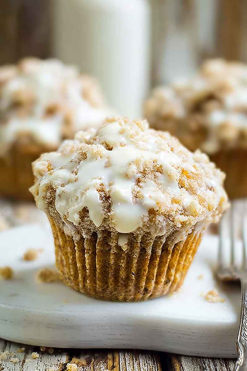 A close-up of a carrot cake muffin with a thick layer of white frosting and a generous sprinkle of crumbly streusel on top.