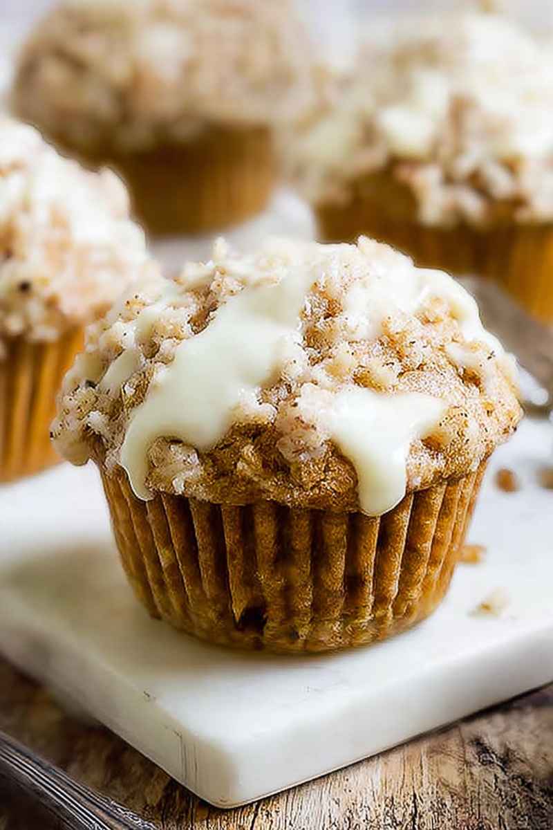 A beautifully presented carrot cake muffin with a glossy white glaze and crumbly streusel topping, placed on a white marble board.