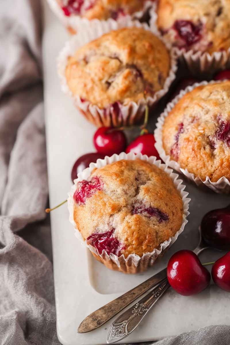 A plate of warm, freshly baked muffins with visible chunks of cherries, accompanied by fresh cherries and a vintage fork.
