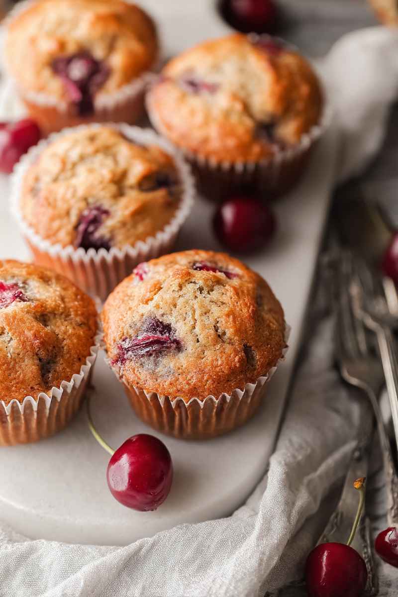 A plate of golden-brown cherry muffins with fresh cherries scattered around, showcasing their homemade appeal.