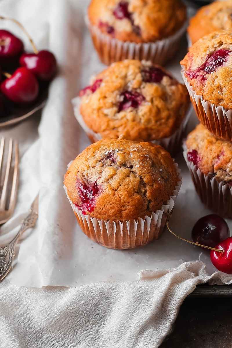 A close-up of golden muffins studded with juicy red cherries, placed on a rustic cloth with additional cherries nearby.