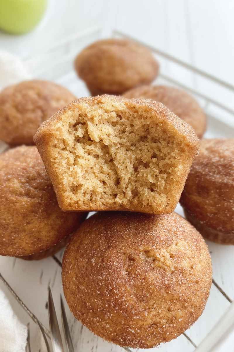 A stack of cinnamon applesauce muffins on a cooling rack, each generously coated in cinnamon sugar, with one muffin cut open to show its fluffy interior.