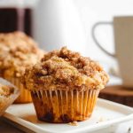 A close-up of a golden-brown cinnamon streusel muffin on a white plate, showcasing its moist texture and crunchy topping.