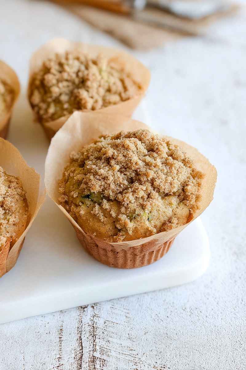 Golden-brown Cinnamon Zucchini Crumb Muffins topped with a rich, buttery crumb topping, sitting on a white marble surface.