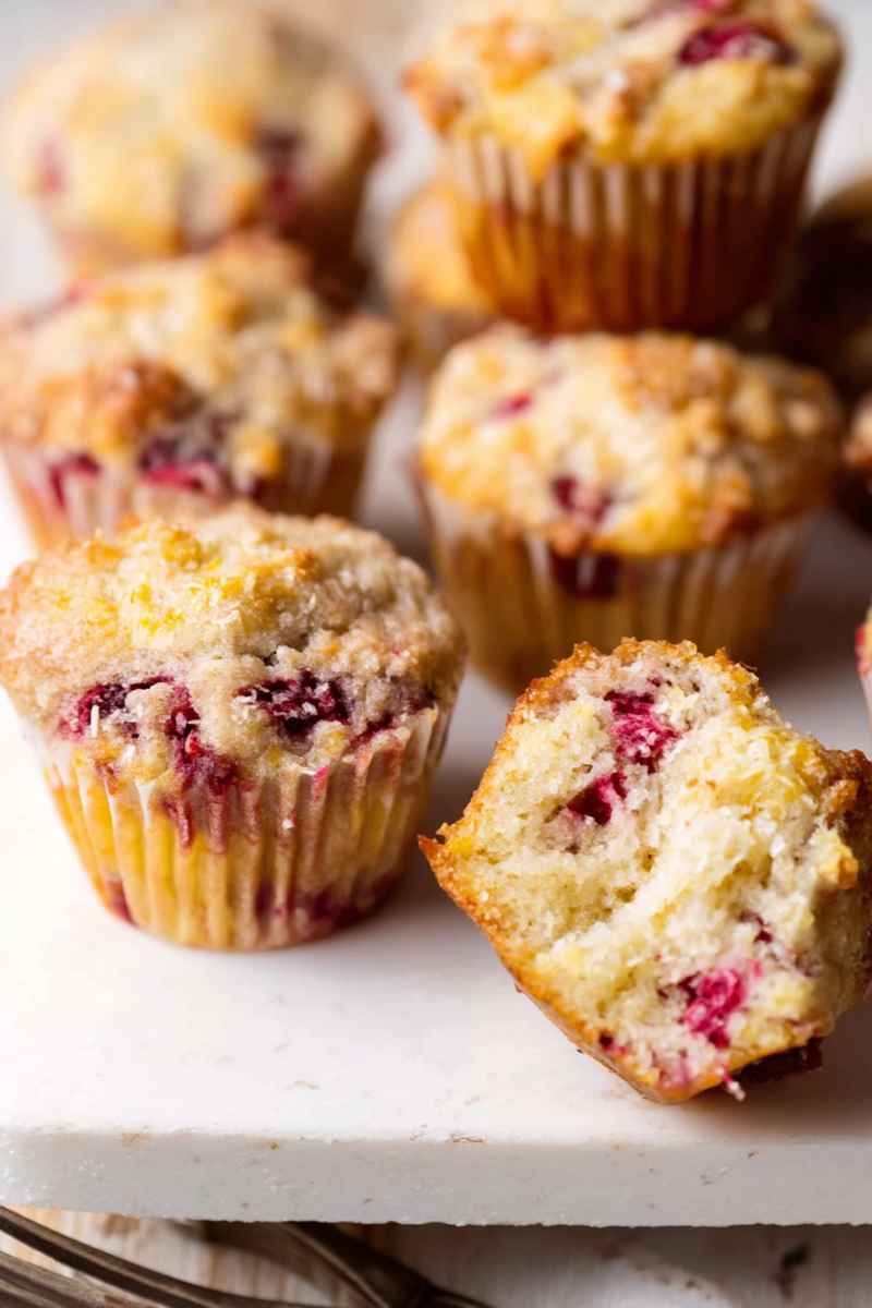 A plate of golden-brown raspberry lemon muffins, some cut open to reveal their moist interior and vibrant red berries.
