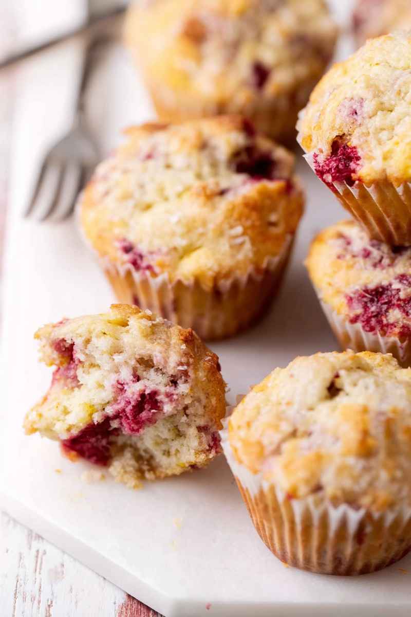 A close-up of freshly baked lemon-raspberry muffins on a white plate, with one split open to show the soft, moist texture and bright red berries.