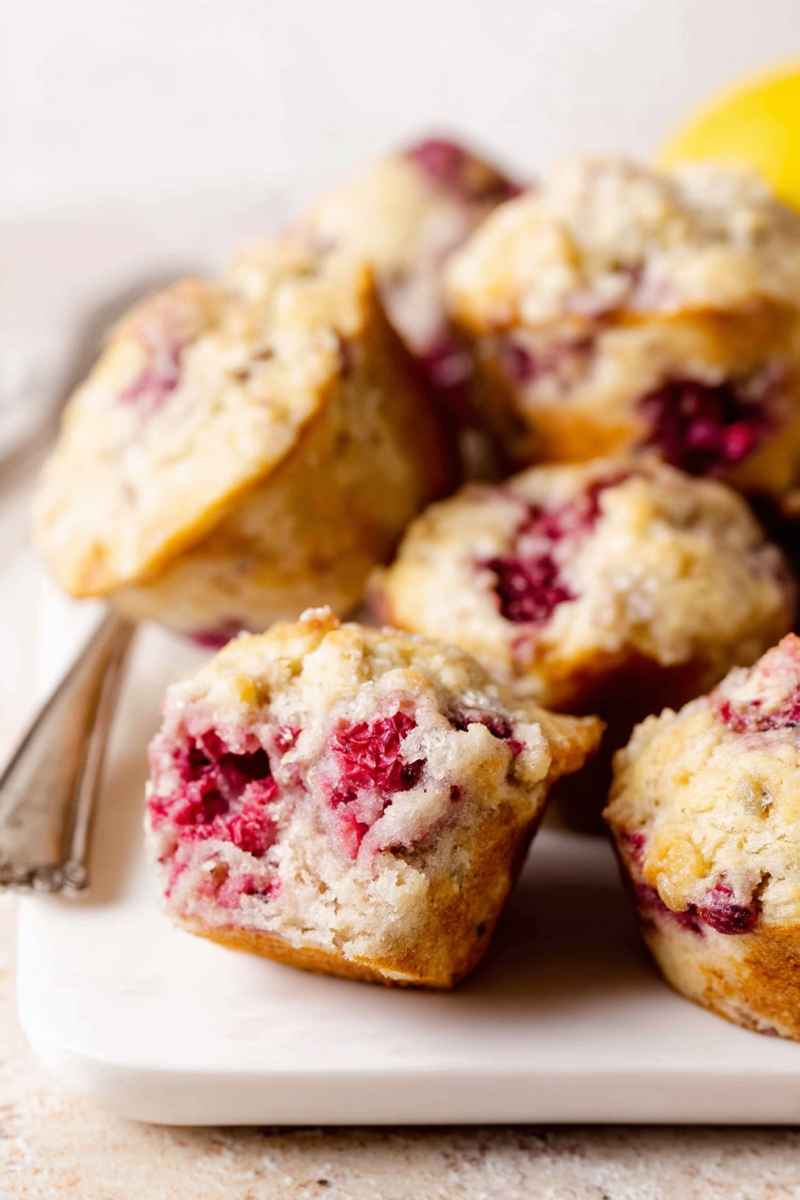 A plate of golden-brown raspberry lemon muffins, some cut open to reveal juicy red berries and a soft, moist interior.