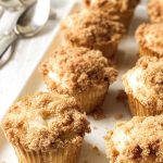 A plate of coffee cake muffins with a creamy filling and a sprinkling of cinnamon sugar crumble, placed near a spoon and FAGE yogurt container.