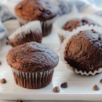 Dark, moist chocolate muffins dusted with powdered sugar, arranged on a white plate with scattered chocolate chips.