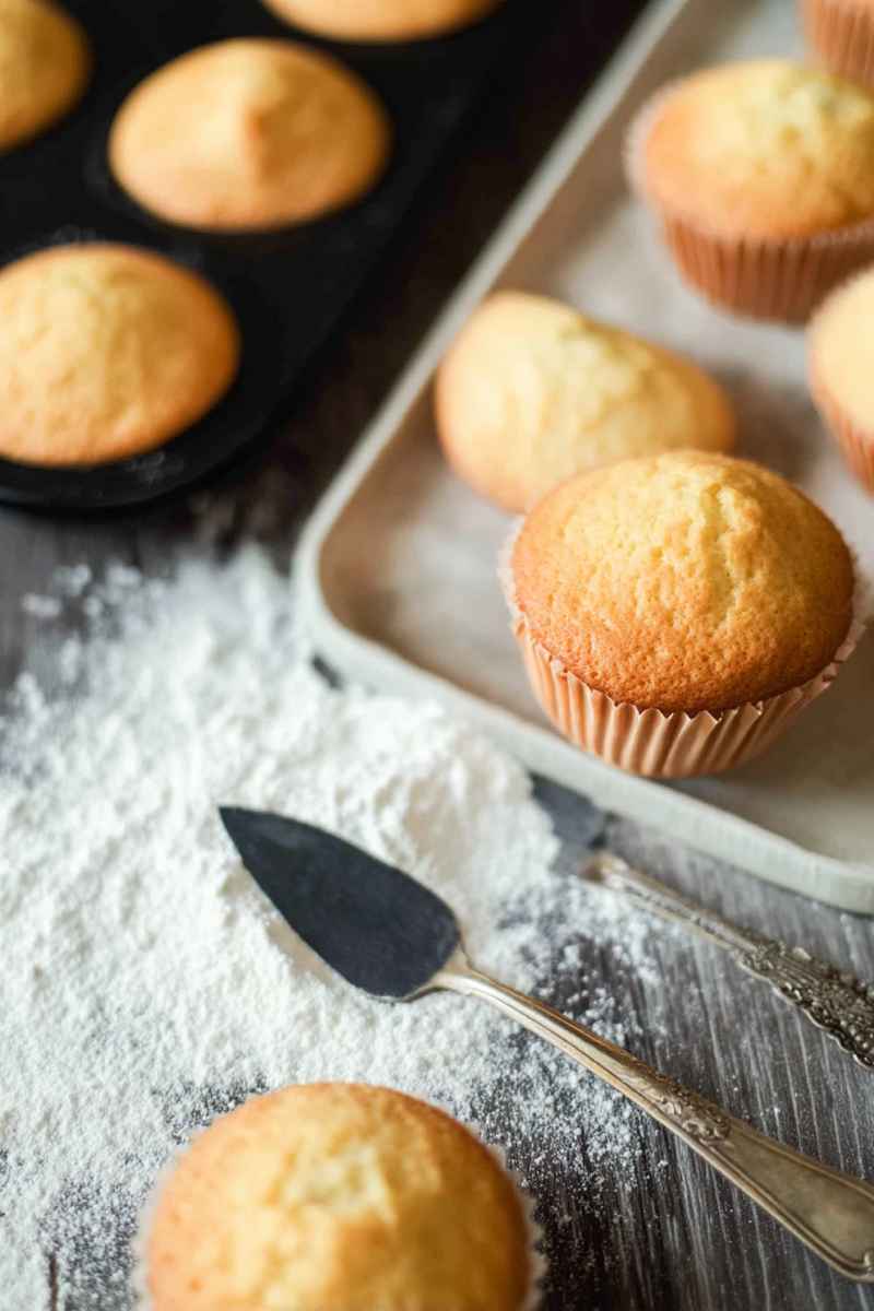 Freshly baked vanilla muffins arranged on a white surface, surrounded by baking tools like spoons and flour.