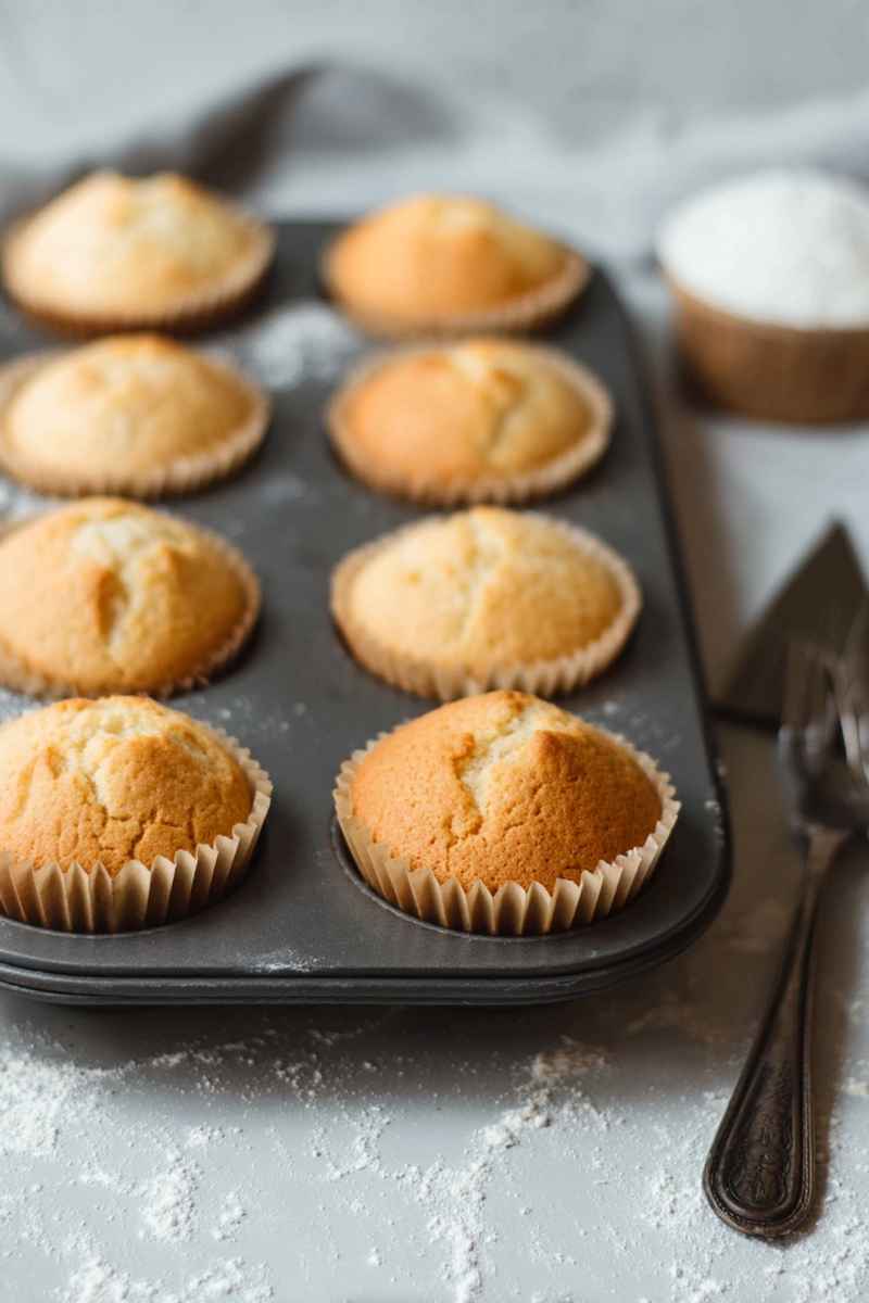 Six golden-brown vanilla muffins in a black muffin tin, showcasing their light and airy texture.