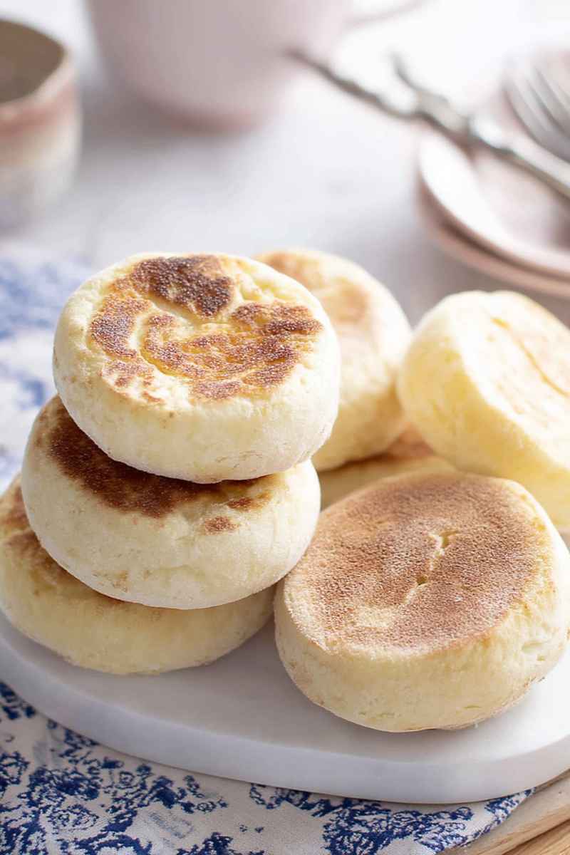 A stack of golden-brown English muffins on a white plate, showcasing their fluffy texture and inviting appearance.