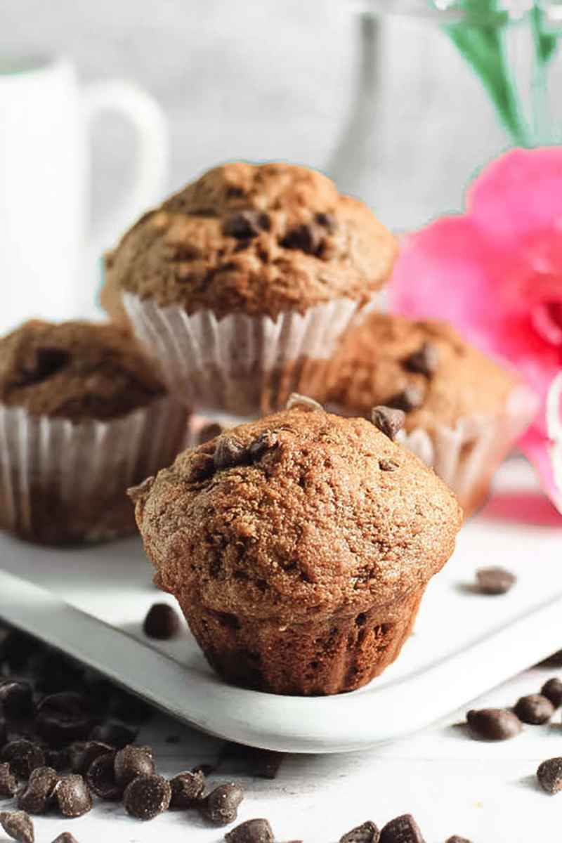 Close-up of Espresso Chocolate Chip Muffins on a white plate, with scattered chocolate chips and pink flowers in the background.