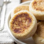 A close-up of gluten-free English muffins stacked on a white plate, highlighting their texture and golden color.