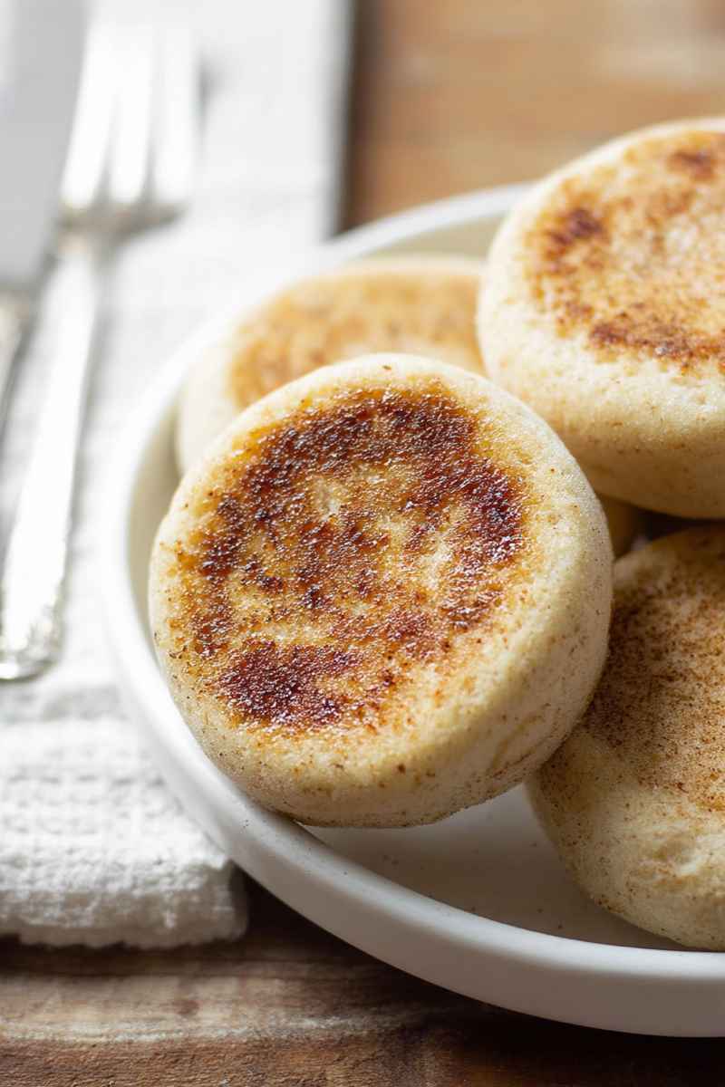 A close-up of gluten-free English muffins stacked on a white plate, highlighting their texture and golden color.