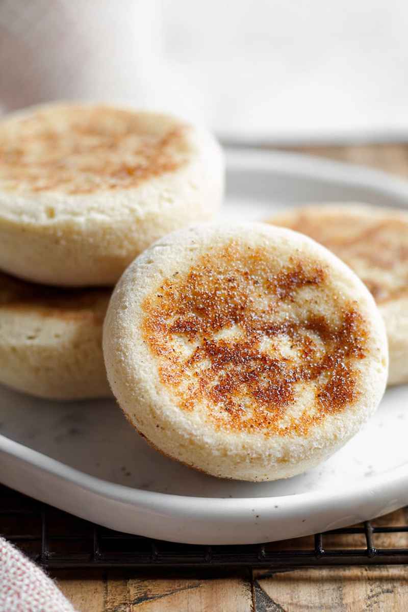 A stack of golden-brown gluten-free English muffins on a white plate, showcasing their characteristic nooks and crannies.