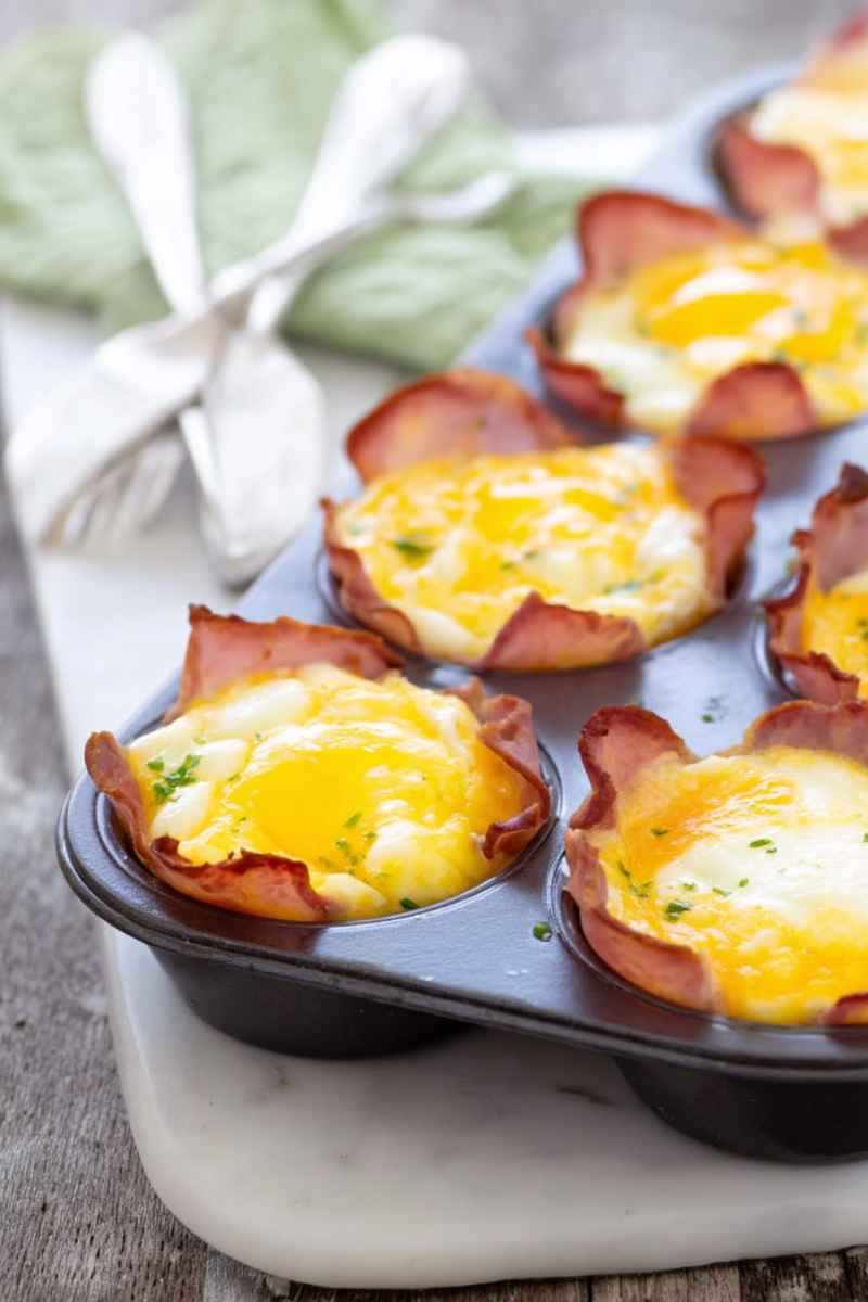 Baked egg cups in a muffin tin lined with ham slices, garnished with fresh herbs.