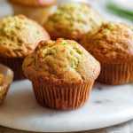 Golden courgette muffins with a sprinkle of green zest, displayed on a rustic table.