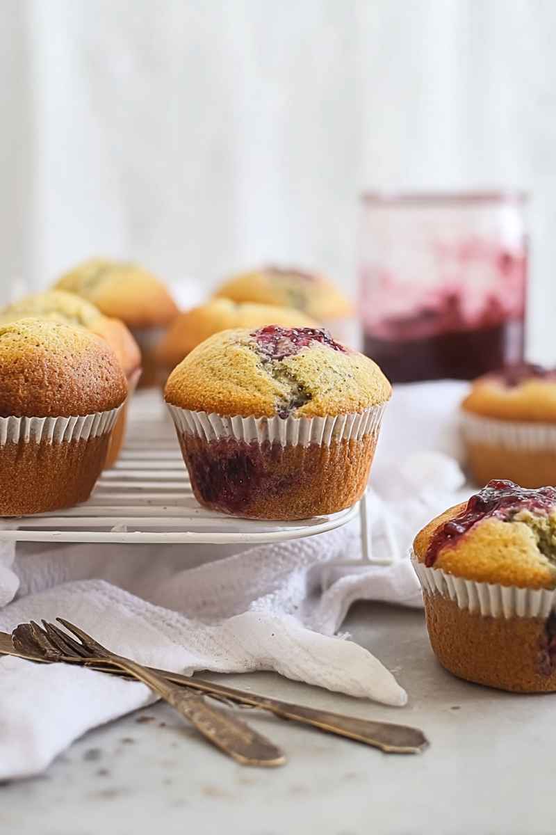 A close-up of golden muffins with visible fruit fillings, arranged on a cooling rack, accompanied by a jar of jam and vintage utensils.