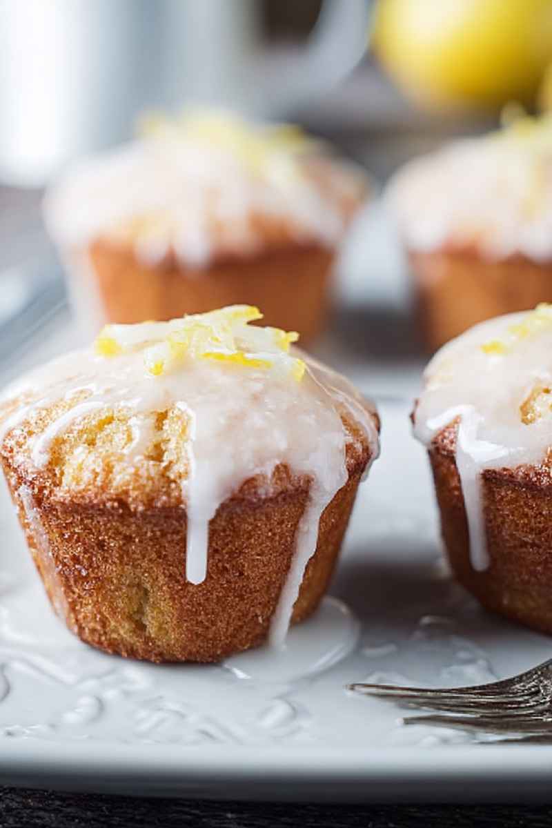 Moist lemon muffins drizzled with a white glaze and garnished with lemon zest, served on a white plate.
