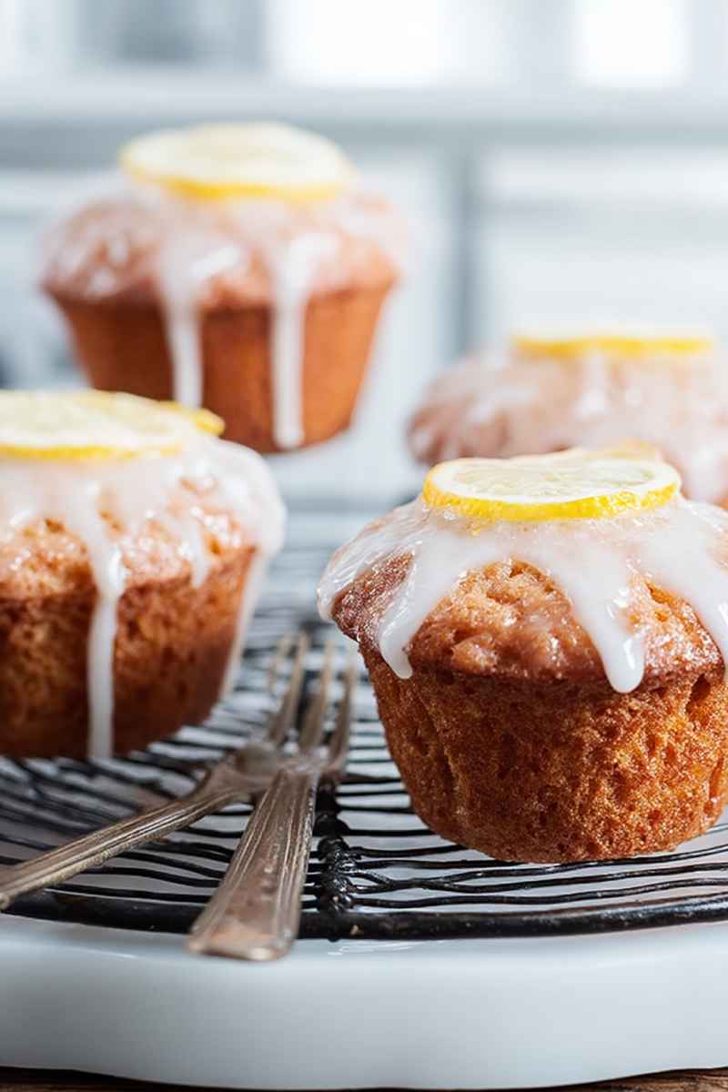 Golden-brown lemon muffins drizzled with white glaze and garnished with lemon slices, arranged on a black-and-white striped cooling rack.