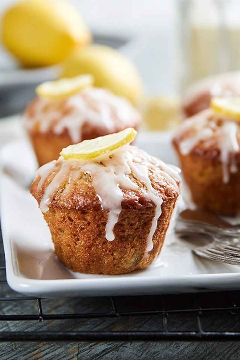 Golden-brown lemon muffins topped with a lemon slice and drizzled with glaze, arranged on a white plate.