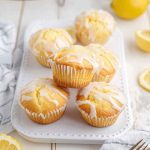A plate of golden Lemon Pound Cake Muffins drizzled with a creamy white glaze, surrounded by fresh lemon slices.
