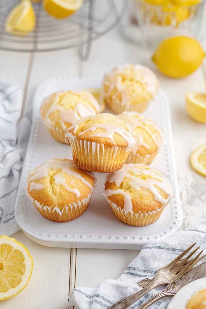 A plate of golden Lemon Pound Cake Muffins drizzled with a creamy white glaze, surrounded by fresh lemon slices.