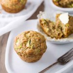A zucchini muffin topped with butter, served on a white plate with a fork nearby.