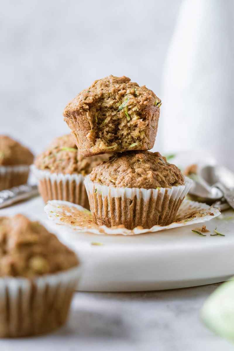Stacked zucchini muffins on a white plate, one cut open to reveal a soft, moist interior.