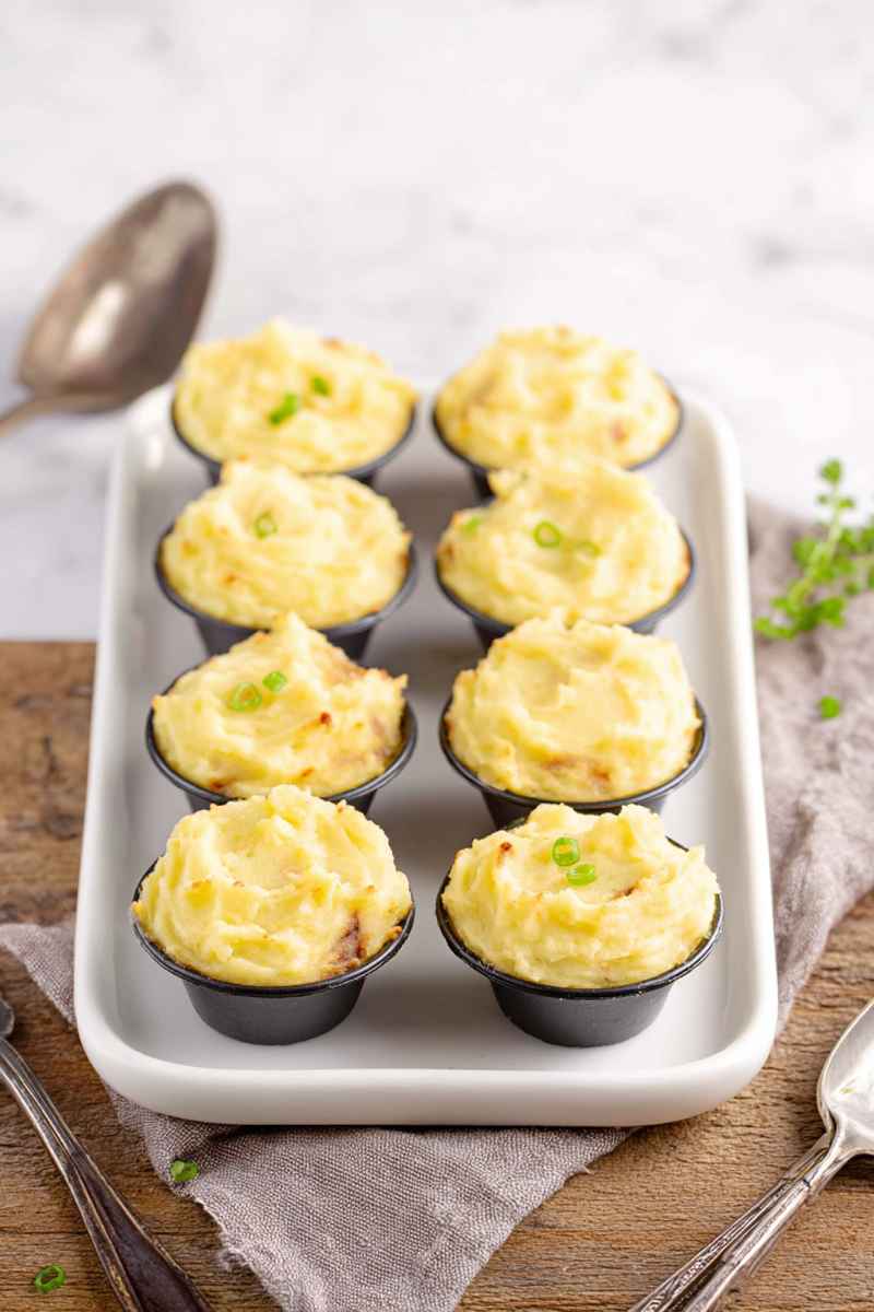 Mini mashed potato cups in black ramekins, garnished with green onions, arranged on a white serving tray.