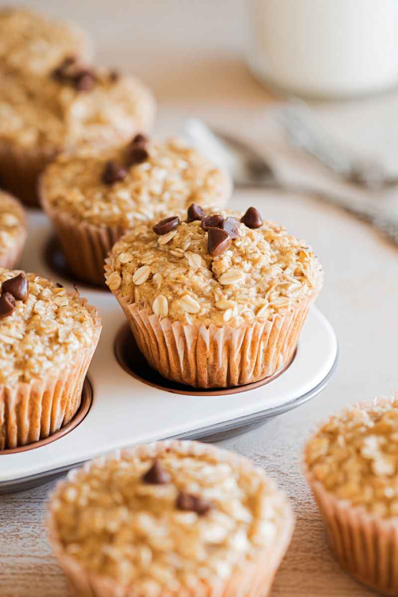 A tray of freshly baked oatmeal muffins with chocolate chips, placed on a wooden surface near a glass of milk.