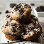 Oreo muffins stacked on a white plate, one cut open to reveal the moist, cookie-filled interior.