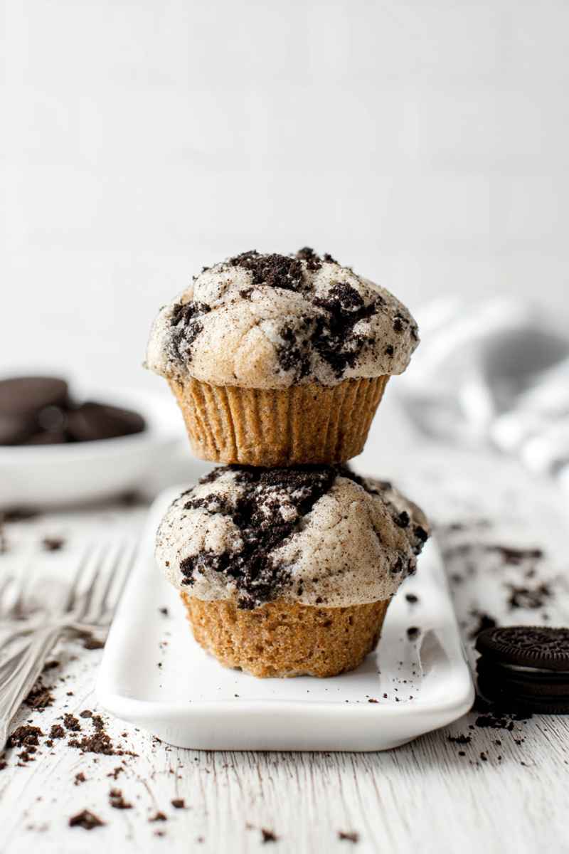 Two Oreo muffins stacked on a white plate, surrounded by whole Oreos and crumbs, ready to be enjoyed.