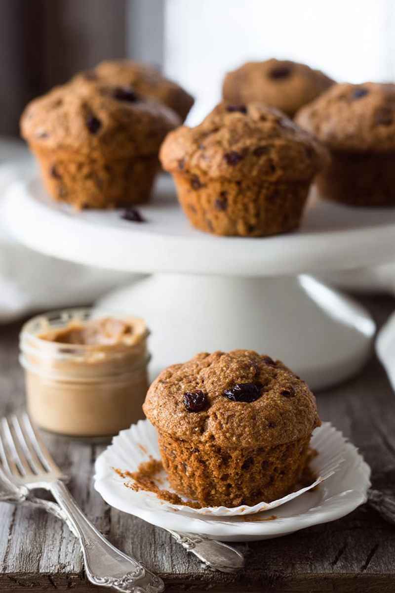 A close-up of a freshly baked Vegan Carrot Raisin Bran Muffin on a white plate, surrounded by more muffins and a jar of peanut butter.
