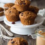 A stack of Vegan Carrot Raisin Bran Muffins on a white cake stand, with scattered raisins and a jar of peanut butter nearby.