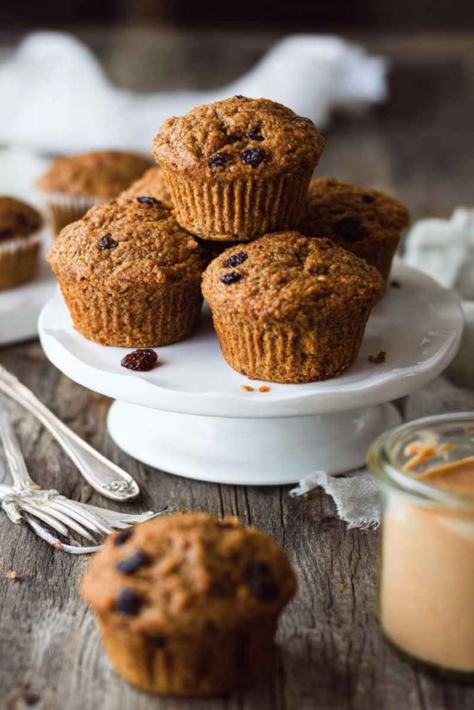 A stack of Vegan Carrot Raisin Bran Muffins on a white cake stand, with scattered raisins and a jar of peanut butter nearby.