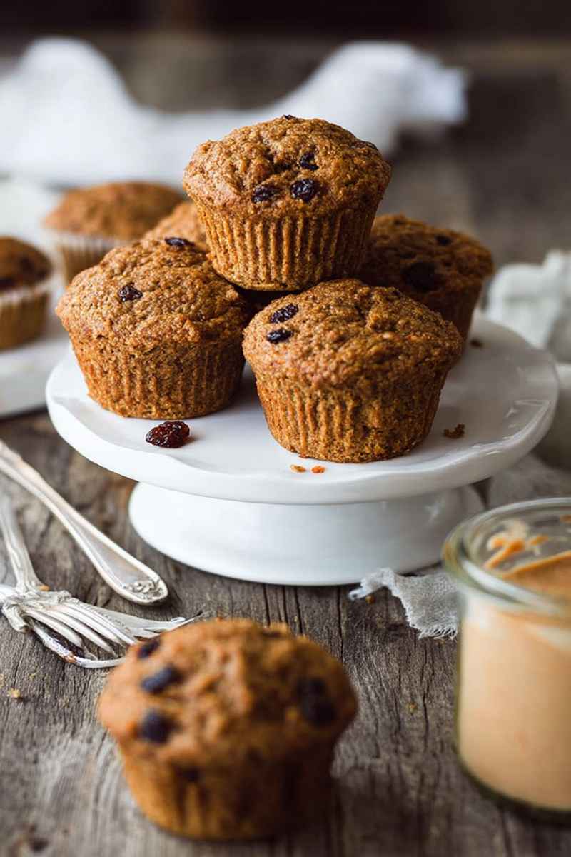 A stack of Vegan Carrot Raisin Bran Muffins on a white cake stand, with scattered raisins and a jar of peanut butter nearby.