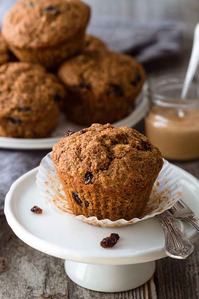 A single Vegan Carrot Raisin Bran Muffin on a white plate, showcasing its moist texture and visible raisins, with a jar of peanut butter in the background.