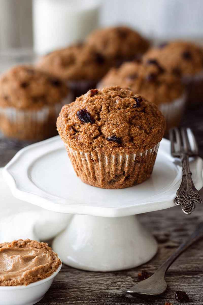 A Vegan Carrot Raisin Bran Muffin on a white cake stand, highlighting its golden-brown exterior and embedded raisins, with a jar of peanut butter and a fork nearby.